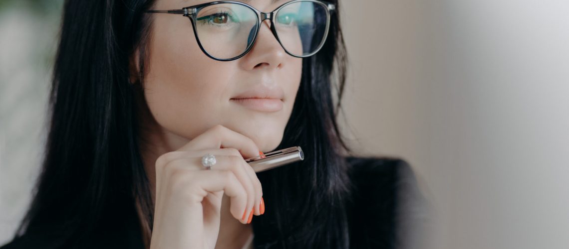 Serious woman author works on new publication, concentrated in display of computer, keeps pen in hand, wears spectacles for eye correction. Ambitious successful female entrepreneur manages business